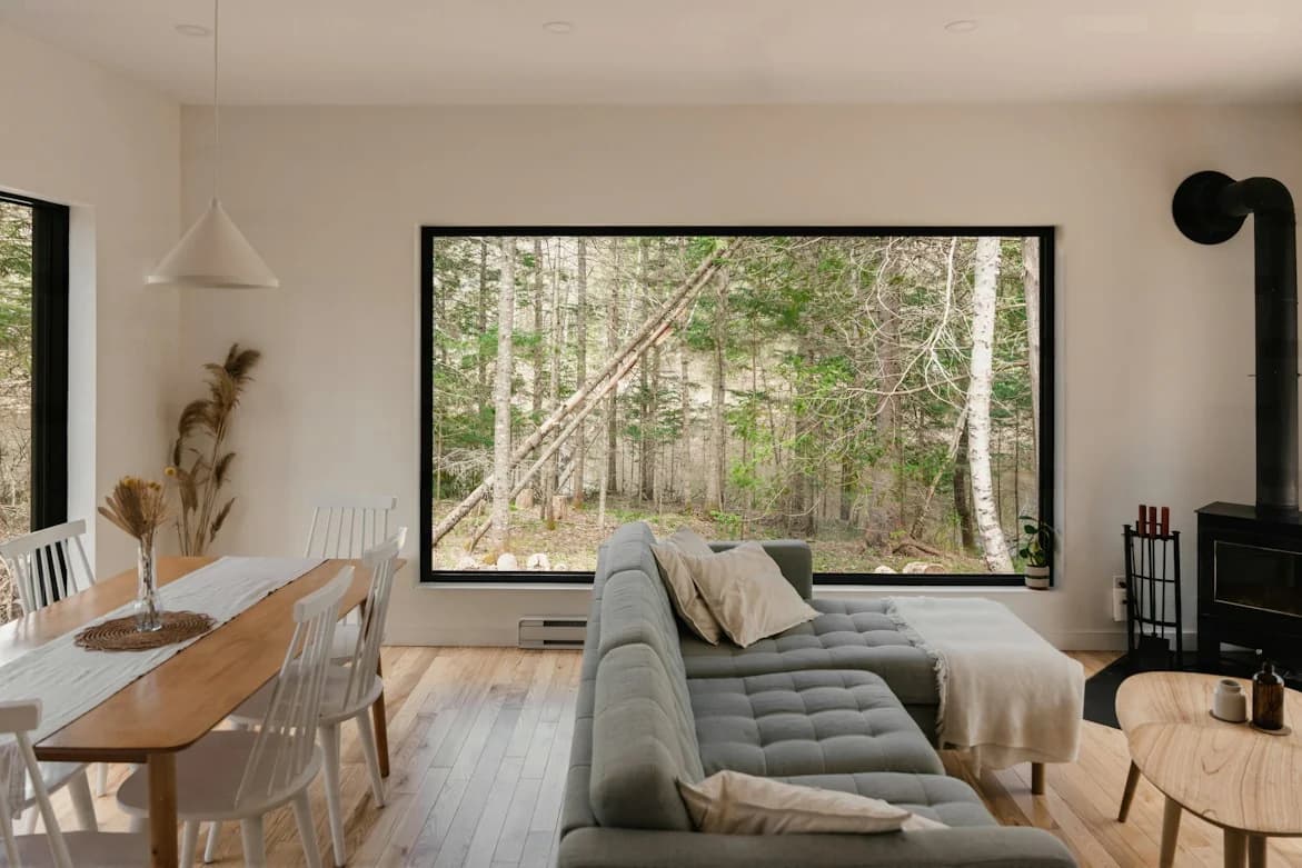 Grey sofa dividing a compact living and dining room, with a timber dining table and a wide picture window facing trees
