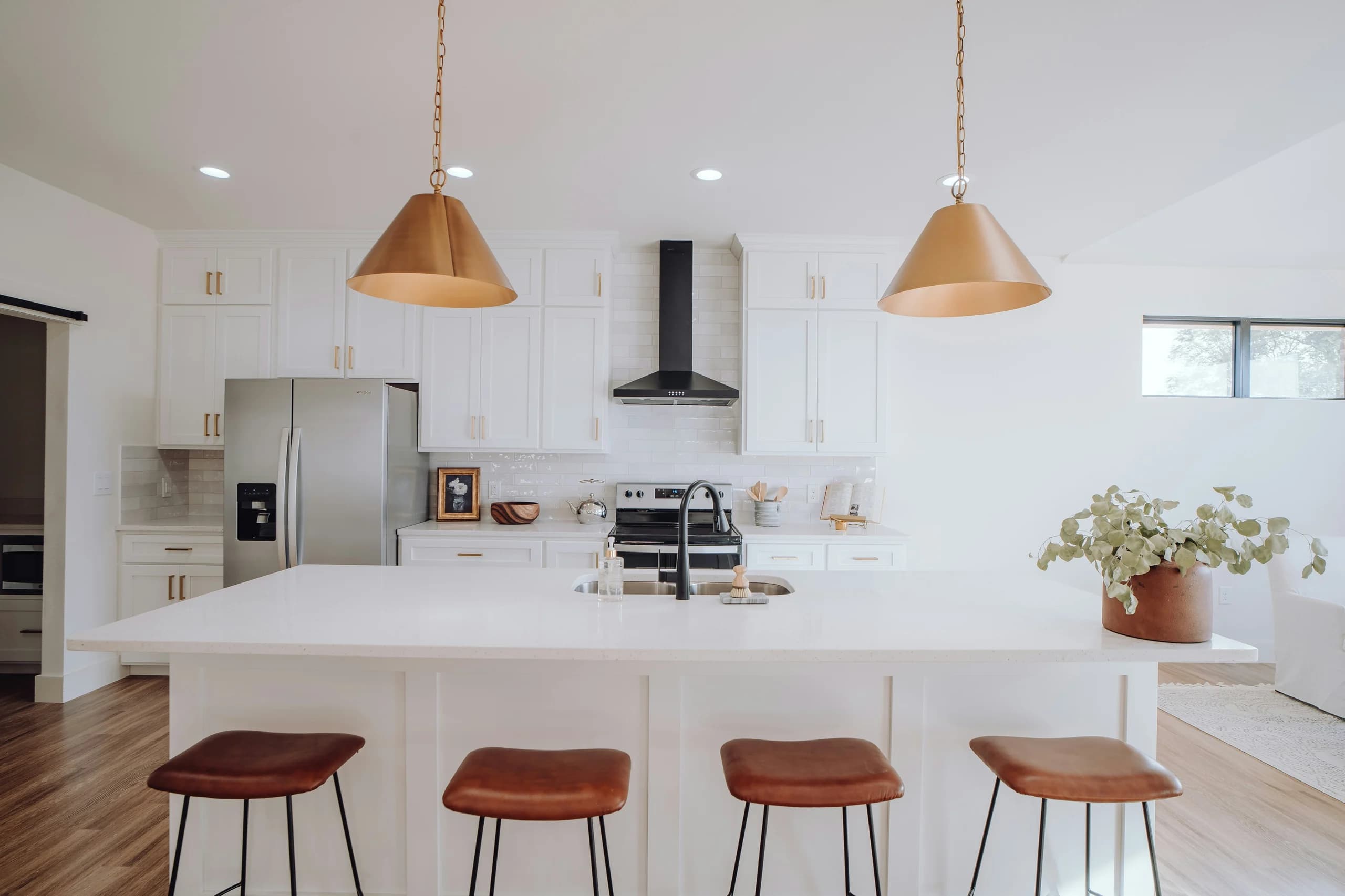 Front view of a bright white kitchen with a large sink island, brown stools, warm metal pendant lights and white cabinetry